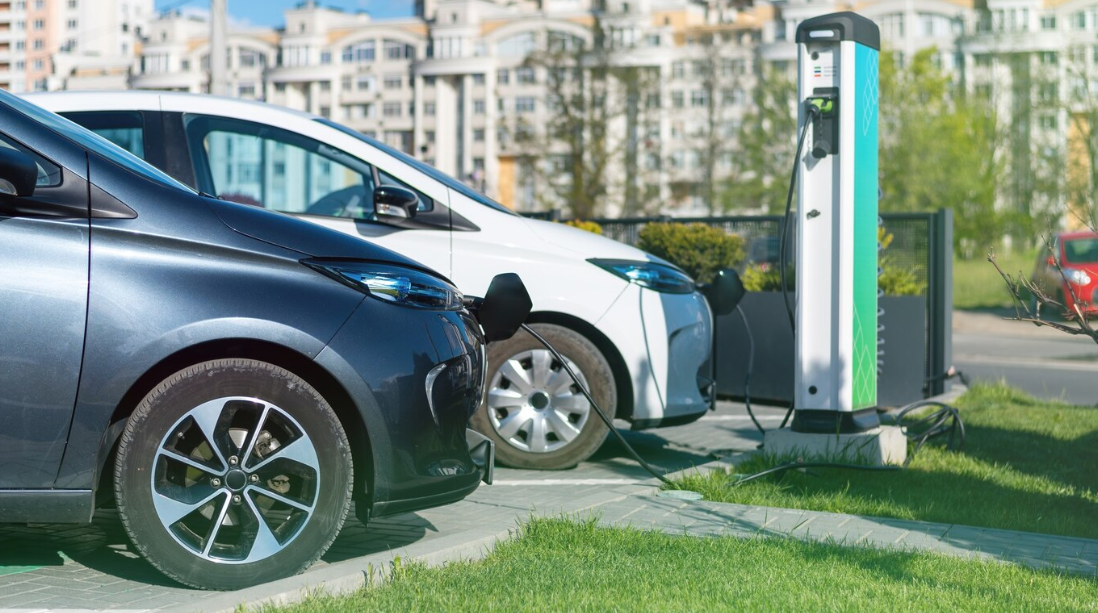 Electric vehicles are charging at a public EV charging station in an urban area with modern apartment buildings in the background.