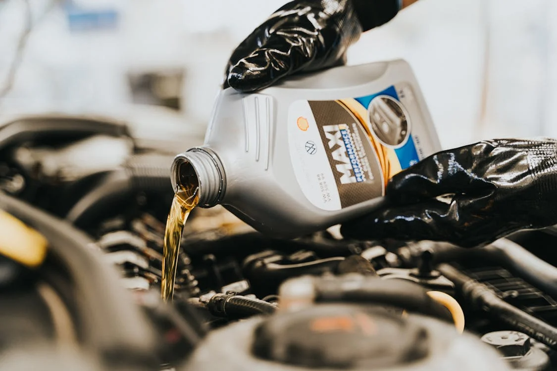 Person wearing black gloves pouring engine oil from a MAXI Performance container into a car engine during a DIY oil change at home.